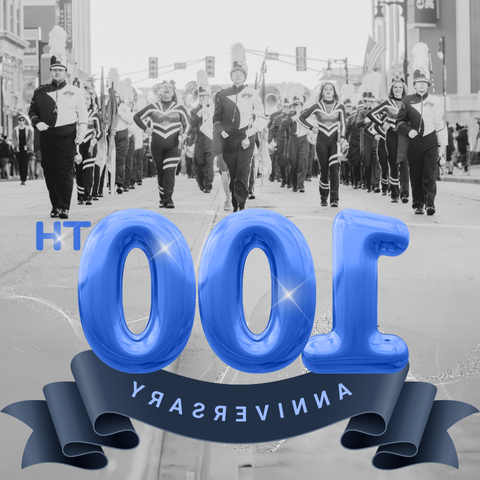 A black and white photo of a marching band parading down a street, led by a drum major. In the foreground, a large blue balloon-style text reads '100th Anniversary' with a blue ribbon banner below it.